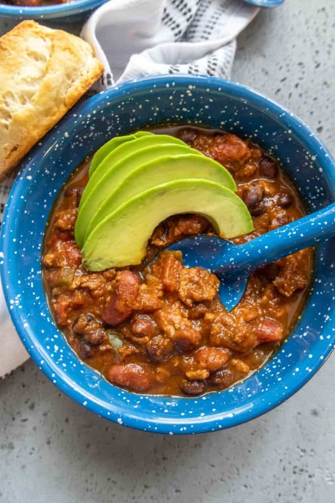 A blue bowl filled with chili garnished with avocado slices and served with a biscuit on the side.