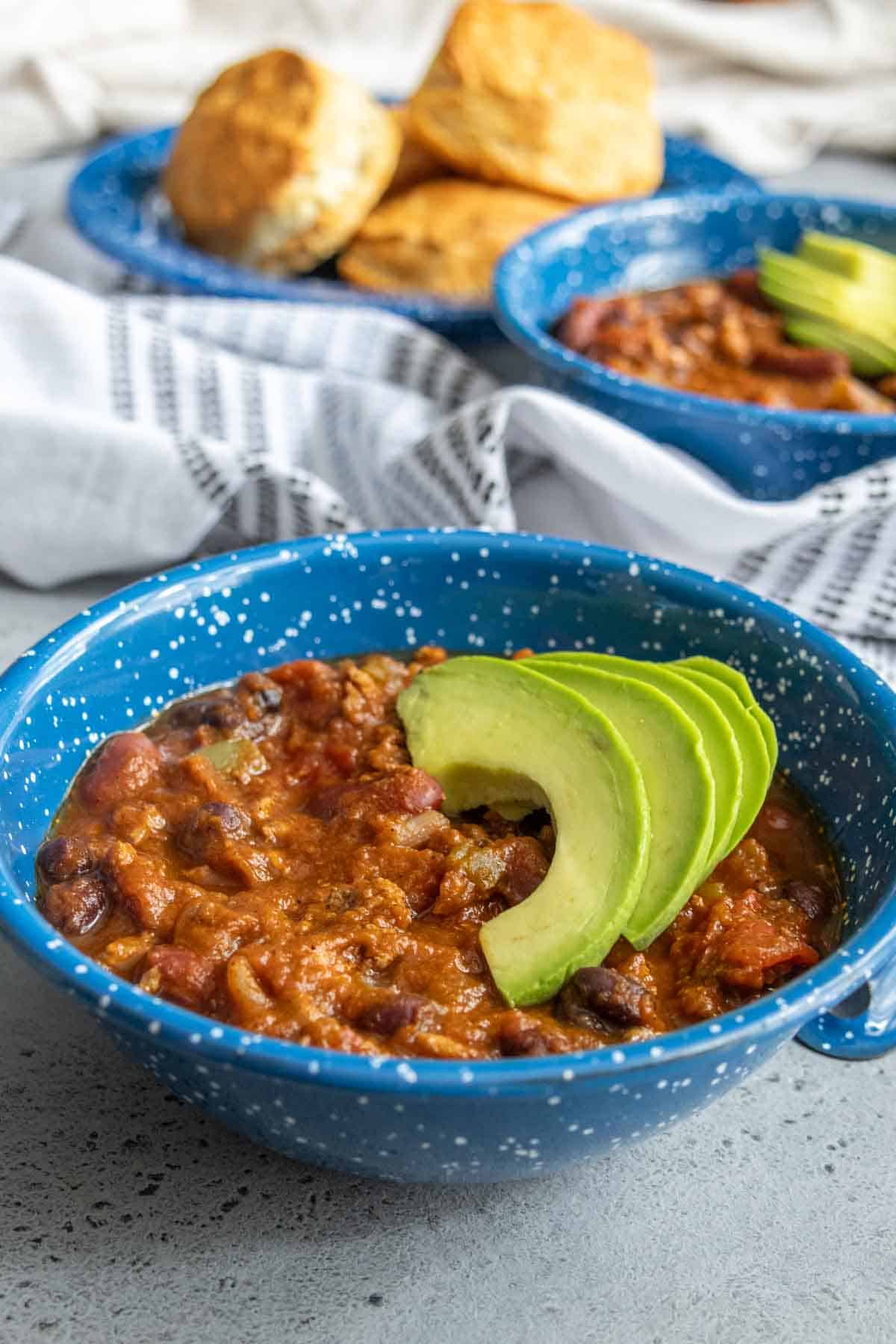 A blue bowl of chili topped with avocado slices, with more chili and biscuits in the background.