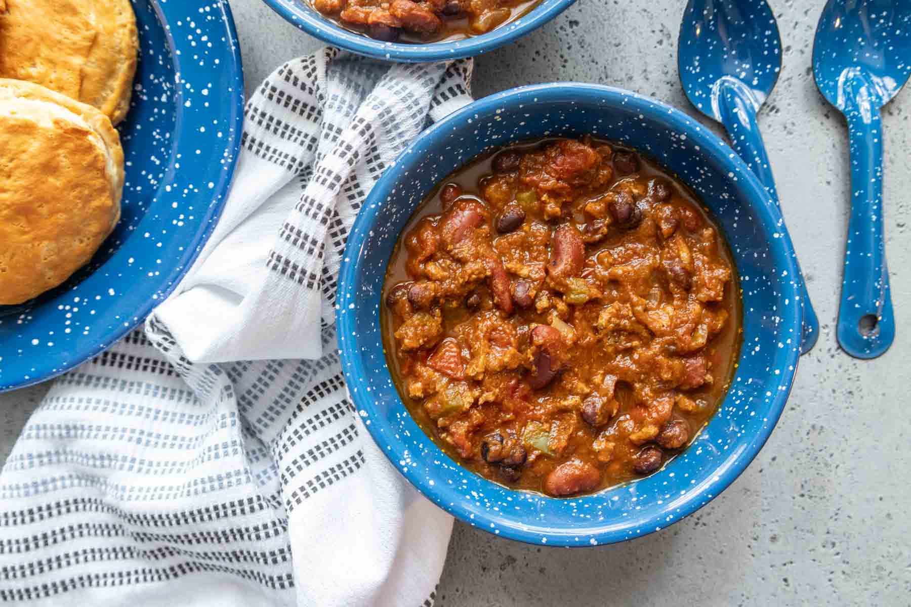 A bowl of pumpkin turkey chili with spoons and biscuits nearby.