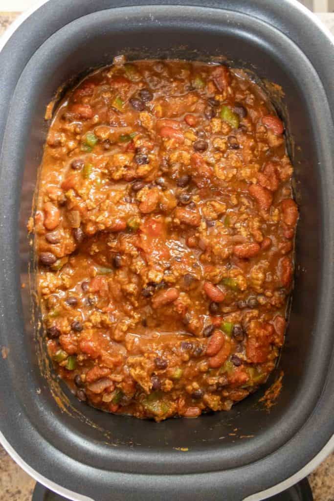 A slow cooker filled with chili, containing beans, diced tomatoes, and ground meat in a thick sauce.