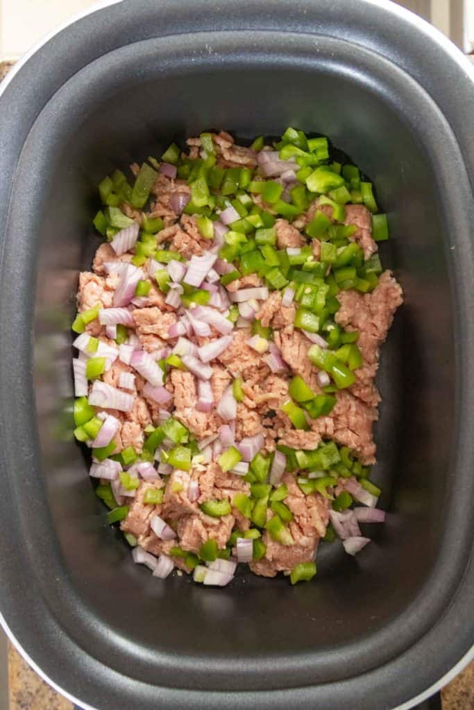 A slow cooker containing raw ground meat mixed with chopped green bell peppers and diced onions.