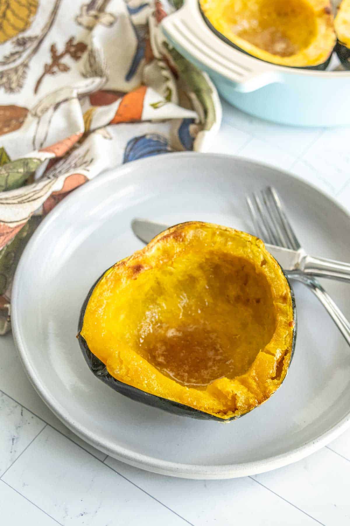 A roasted half of an acorn squash is placed on a gray plate with a silver fork and knife beside it. A colorful napkin and another dish with squash are in the background.