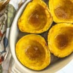 Four baked acorn squash halves with a honey glaze are arranged on a white oval dish. A patterned cloth napkin is placed beside the dish.