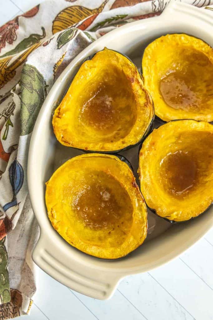 Four baked acorn squash halves with a honey glaze are arranged on a white oval dish. A patterned cloth napkin is placed beside the dish.