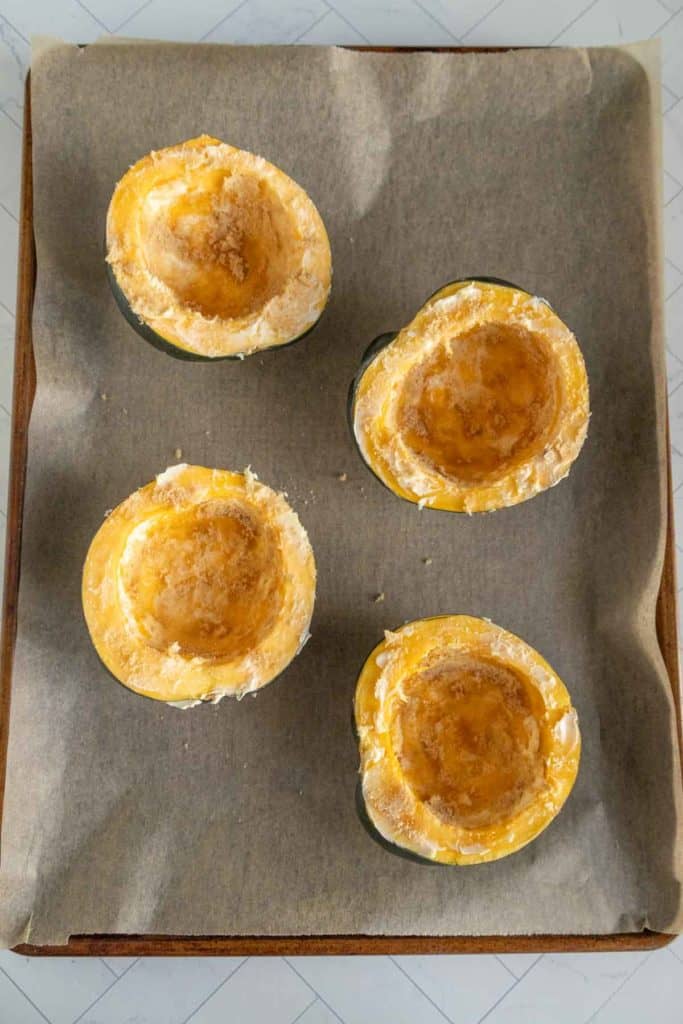 Four halved butternut squash pieces with seasoned tops sit on a parchment-lined baking sheet, ready to be roasted.