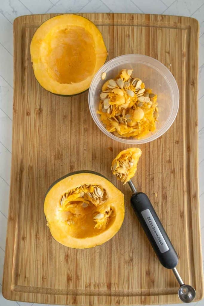 Two halved acorn squash on a wooden cutting board. One half is hollowed out, with a melon baller tool and seeds placed in a plastic container.