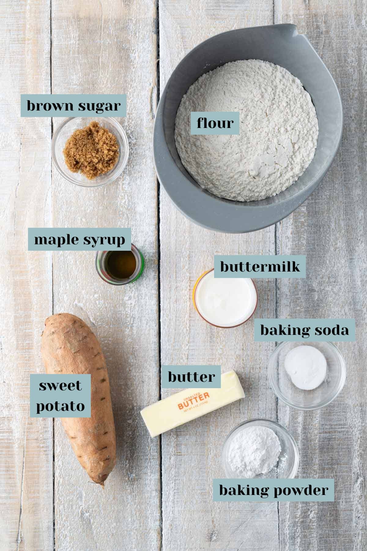 Overhead view of baking ingredients including flour, buttermilk, sweet potato, butter, brown sugar, maple syrup, baking soda, and baking powder on a wooden surface. Each item is labeled with text.