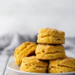 A plate with a stack of five golden-brown biscuits on a light wooden surface, with a blurred background.