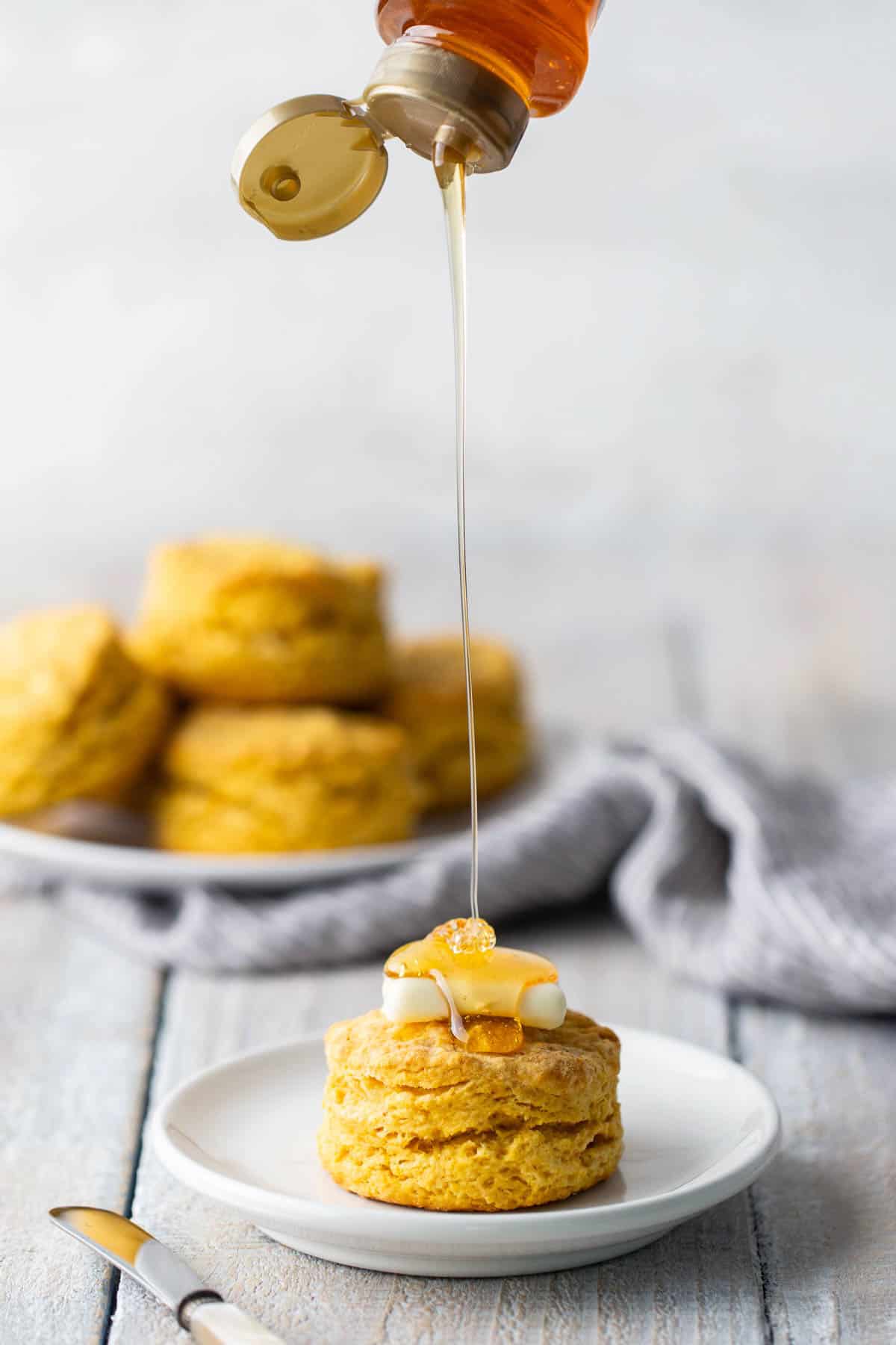 Honey being drizzled on a biscuit placed on a white plate, with additional biscuits and a cloth in the background. A knife rests on the table beside the plate.