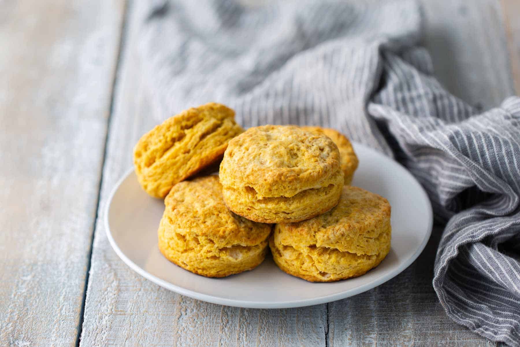 A white plate with five freshly baked biscuits is placed on a wooden surface next to a gray-striped cloth.