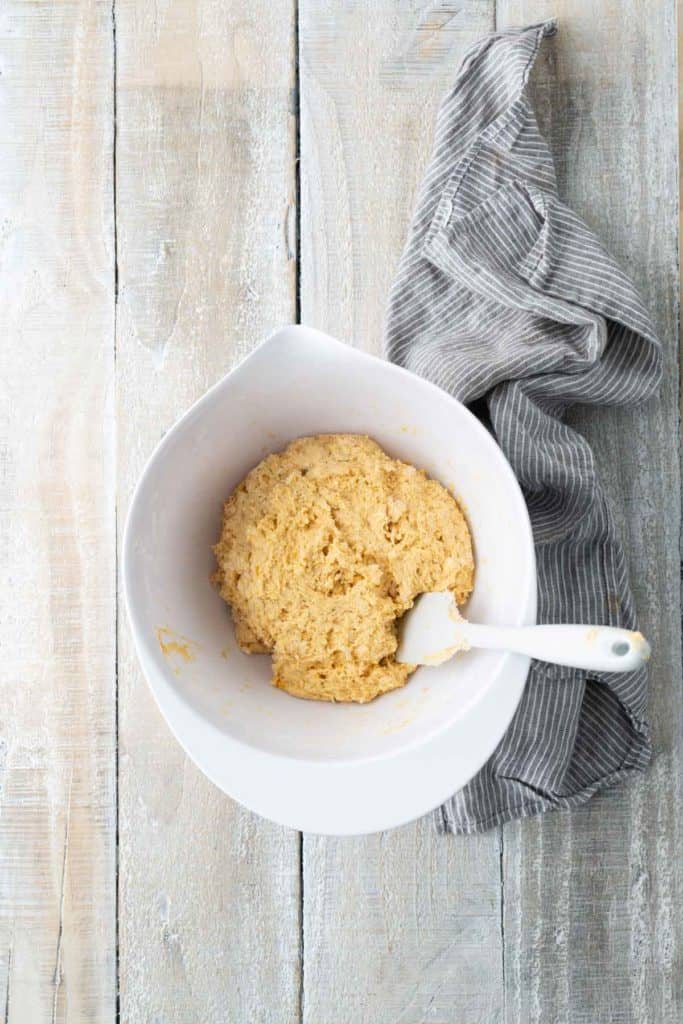 A white mixing bowl on a wooden surface contains cookie dough and a white spatula; a gray striped cloth is beside the bowl.