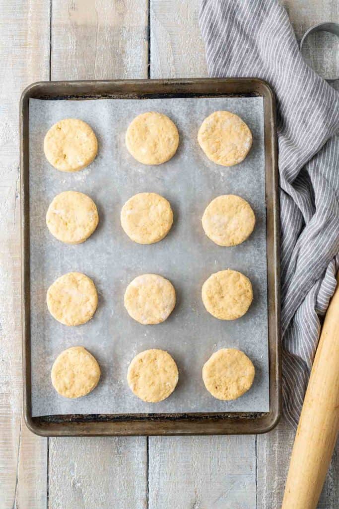 A baking tray with twelve unbaked biscuits arranged in a 3x4 grid on parchment paper. A rolling pin and a striped kitchen towel are next to the tray on a wooden surface.