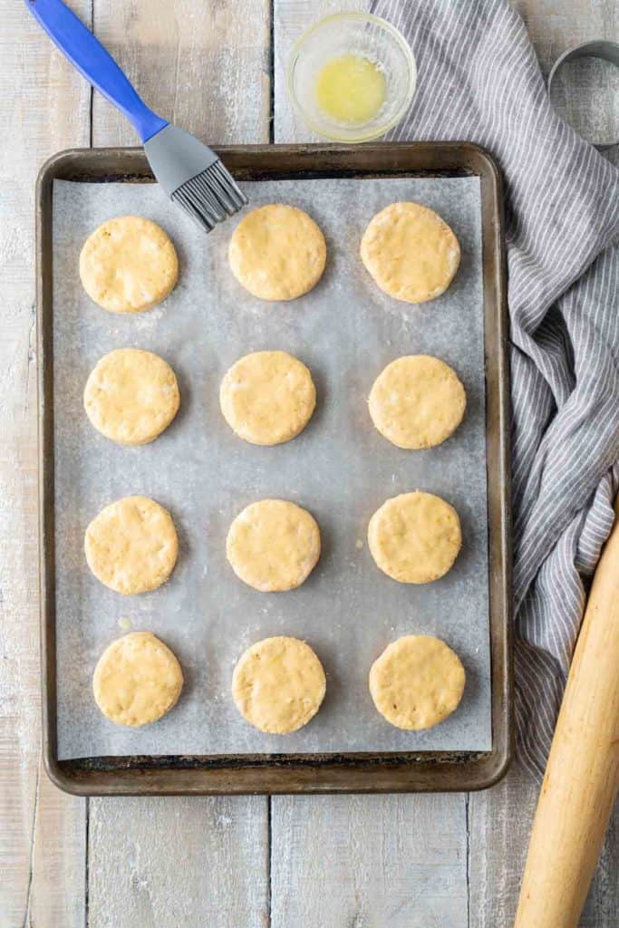 A baking tray lined with parchment paper holds twelve uncooked dough circles. A pastry brush in a bowl with a light liquid, a striped cloth, and a wooden rolling pin are on the table beside the tray.