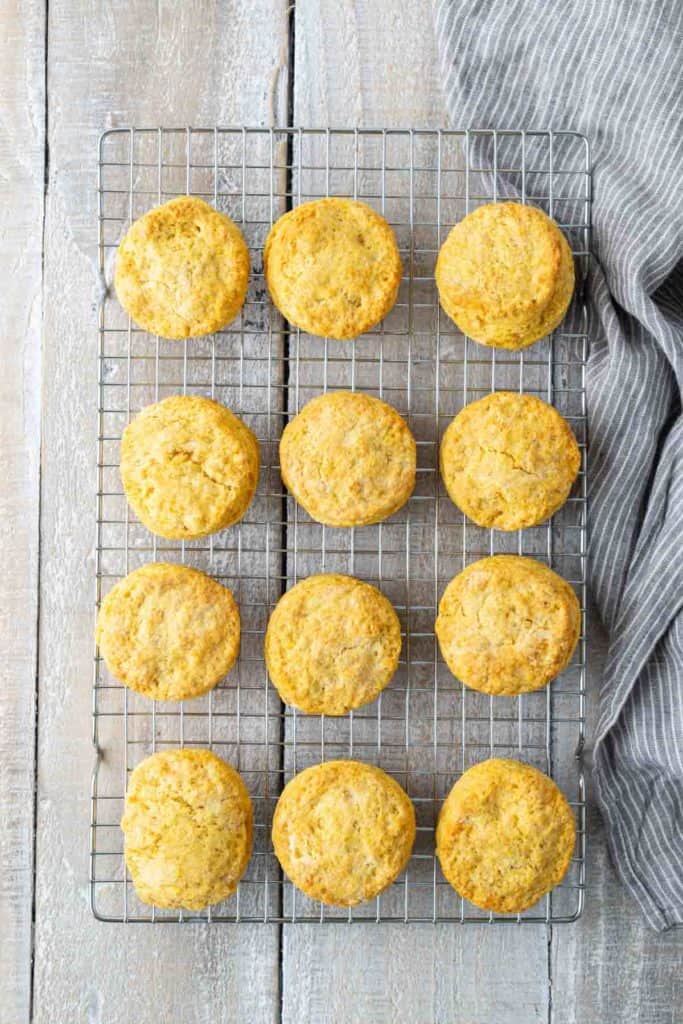 A cooling rack with twelve evenly spaced golden brown biscuits on a wooden surface. A striped cloth is partially visible on the right side.