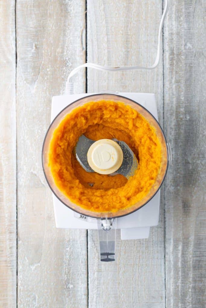 A food processor containing bright orange, blended pumpkin puree on a wooden surface.