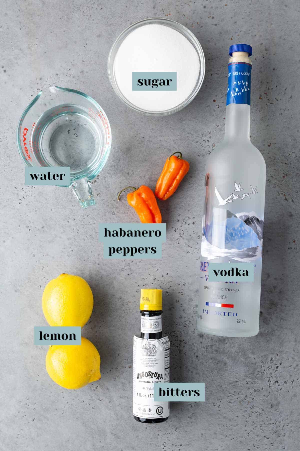 Ingredients for a cocktail are laid out on a table: sugar, water, habanero peppers, a bottle of vodka, lemons, and bitters.