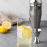 A glass of lemonade with ice cubes and a lemon slice, placed on a gray surface with a stainless steel cocktail shaker and a jigger in the background.