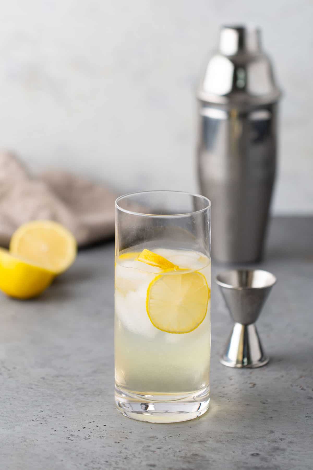 A glass of lemonade with ice cubes and a lemon slice, placed on a gray surface with a stainless steel cocktail shaker and a jigger in the background.