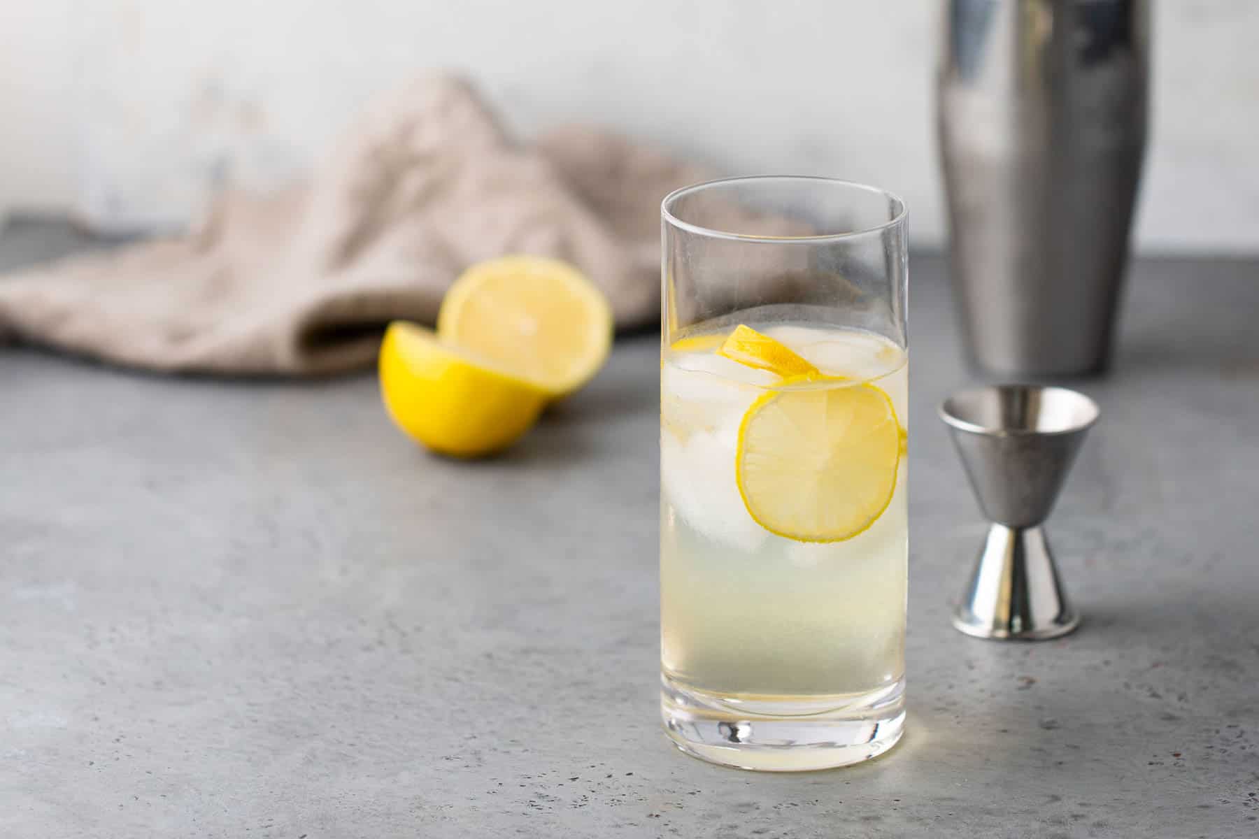 A tall glass of lemonade with ice and a lemon slice, placed on a gray surface. A halved lemon, a small jigger, and a shaker are in the background.