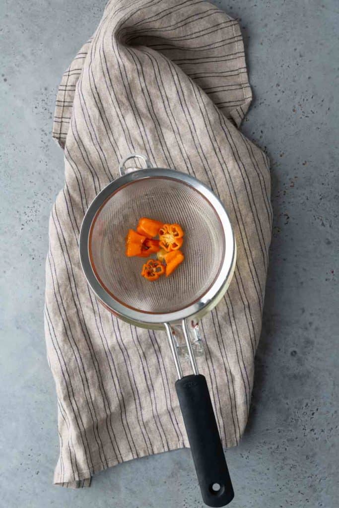 A metal strainer with sliced orange habanero peppers sits on a striped cloth on a gray surface.