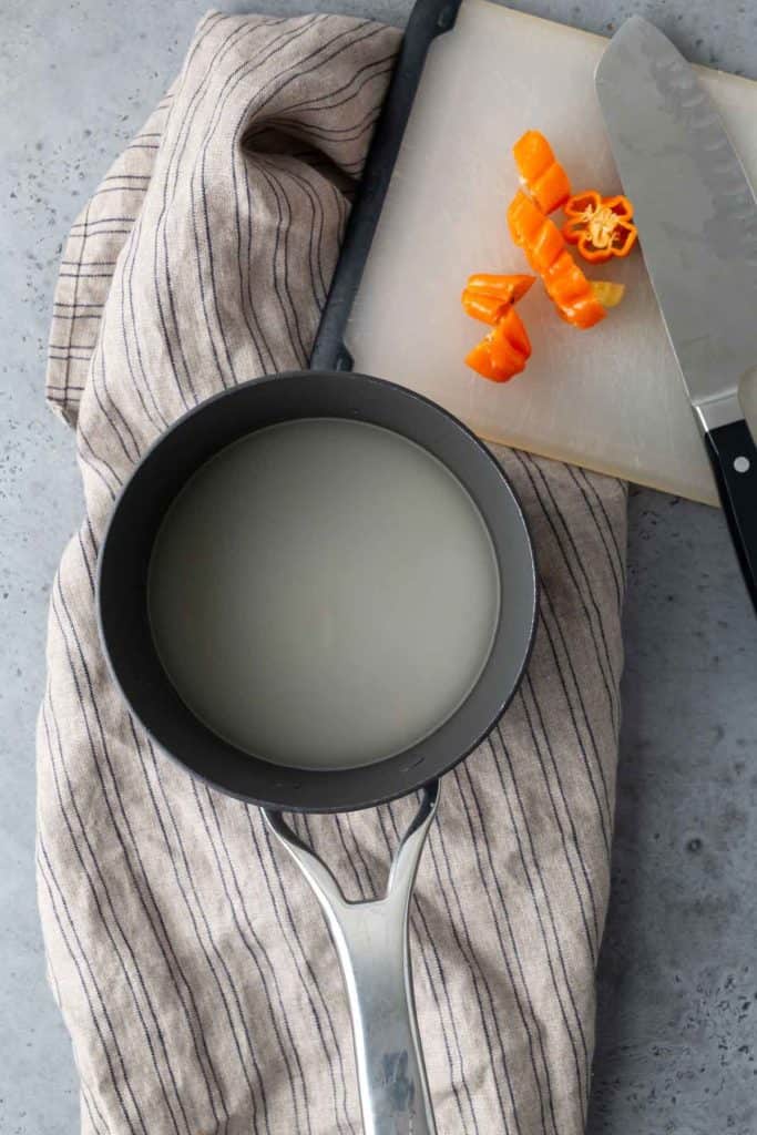 A saucepan containing liquid sits on a striped cloth next to a cutting board with chopped orange peppers and a knife.