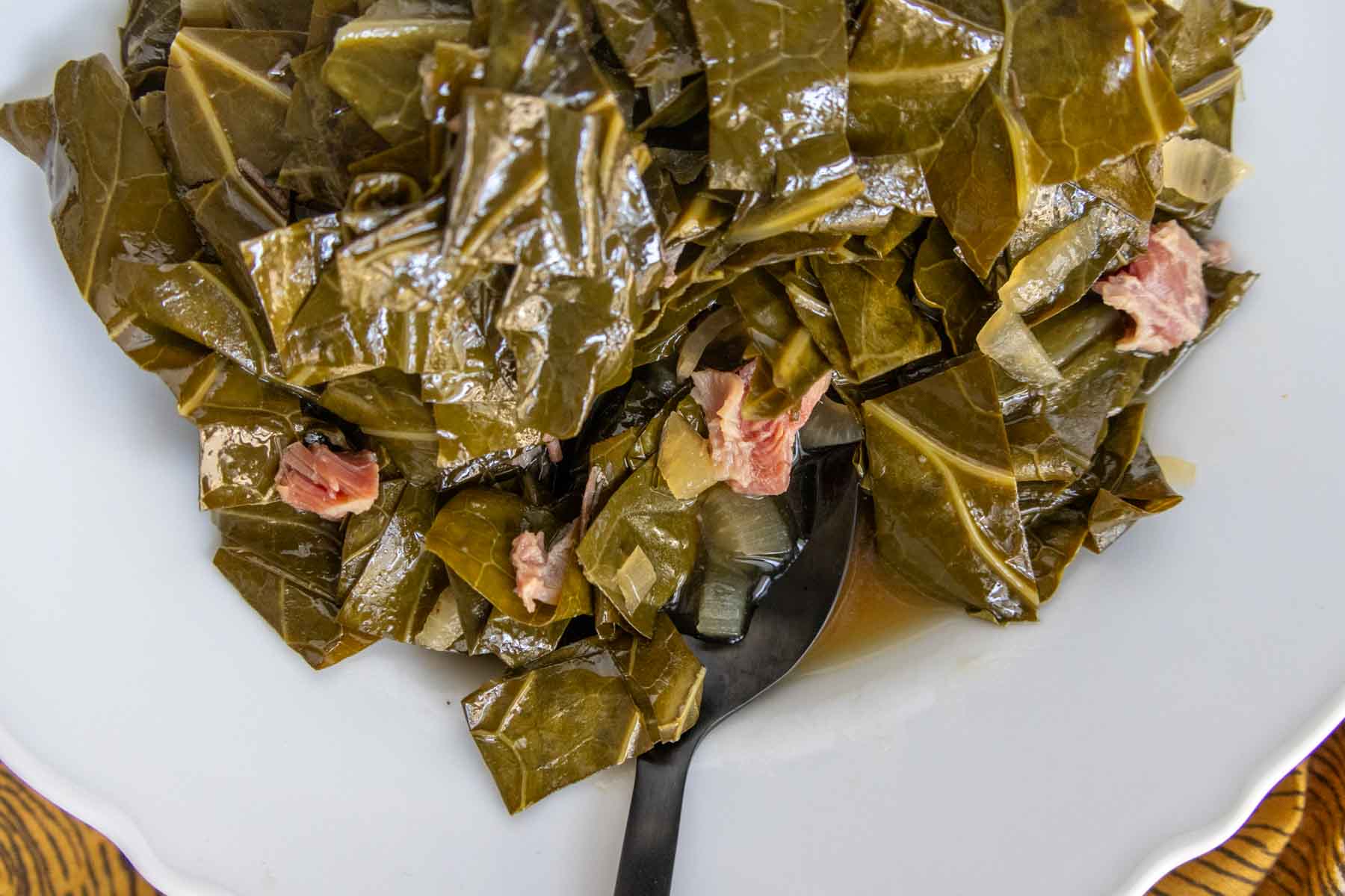 A white bowl containing cooked collard greens mixed with pieces of meat and a black spoon.