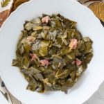 A bowl of cooked collard greens with pieces of meat, served on a patterned cloth, and a black serving spoon nearby.