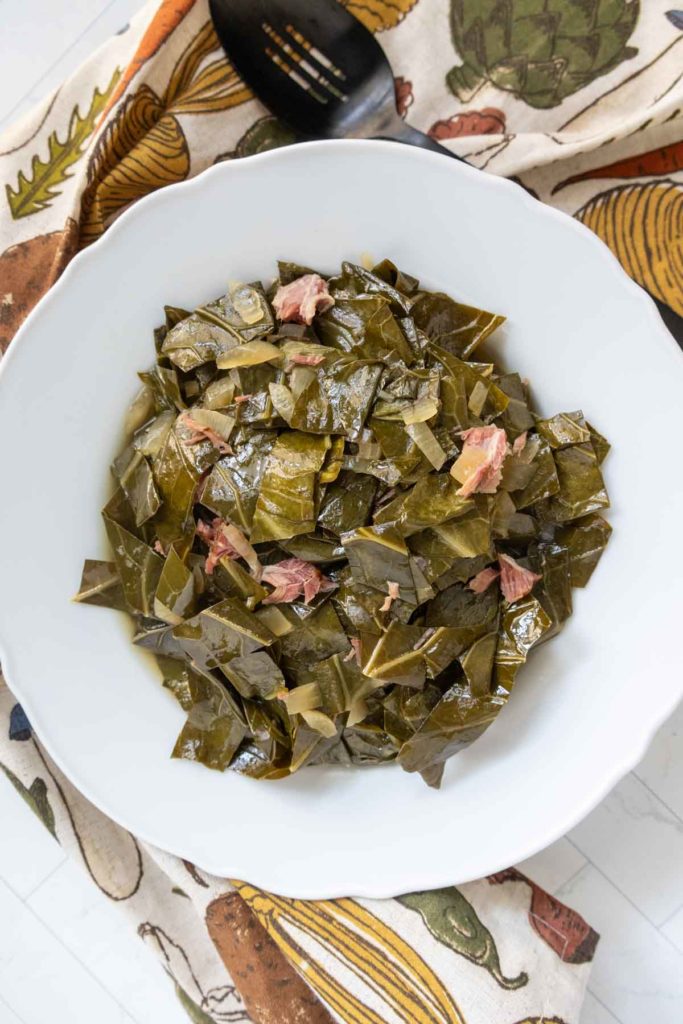 A bowl of cooked collard greens with pieces of meat, served on a patterned cloth, and a black serving spoon nearby.