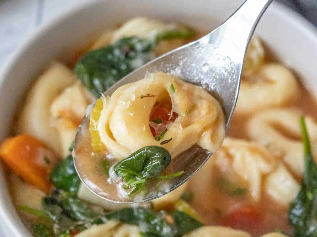 Spoonful of tortellini soup held over bowl.