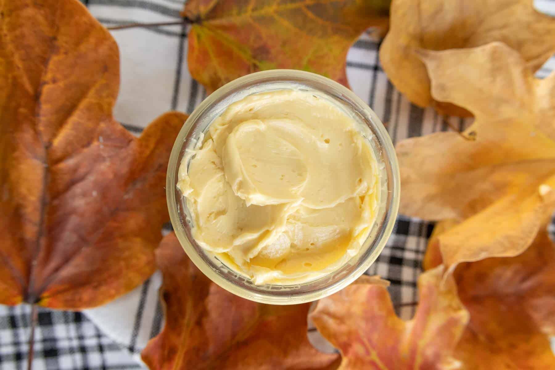 A glass jar filled with creamy butter, surrounded by autumn leaves on a plaid fabric background.