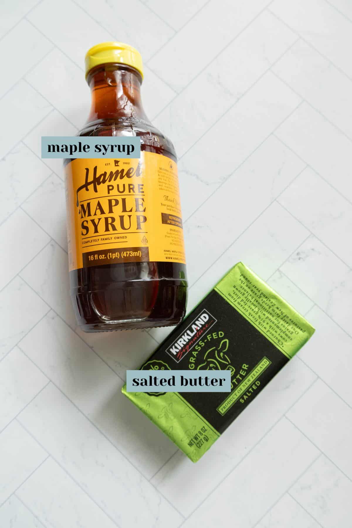 Bottle of maple syrup and a pack of salted butter on a light surface.