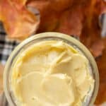 A jar of creamy butter sits on a surface with autumn leaves around it.