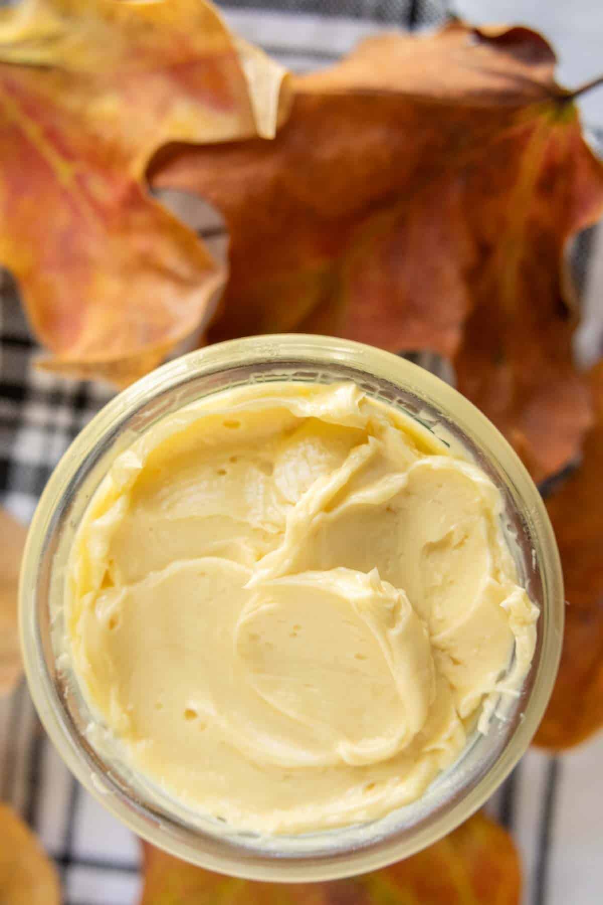 A jar of creamy butter sits on a surface with autumn leaves around it.