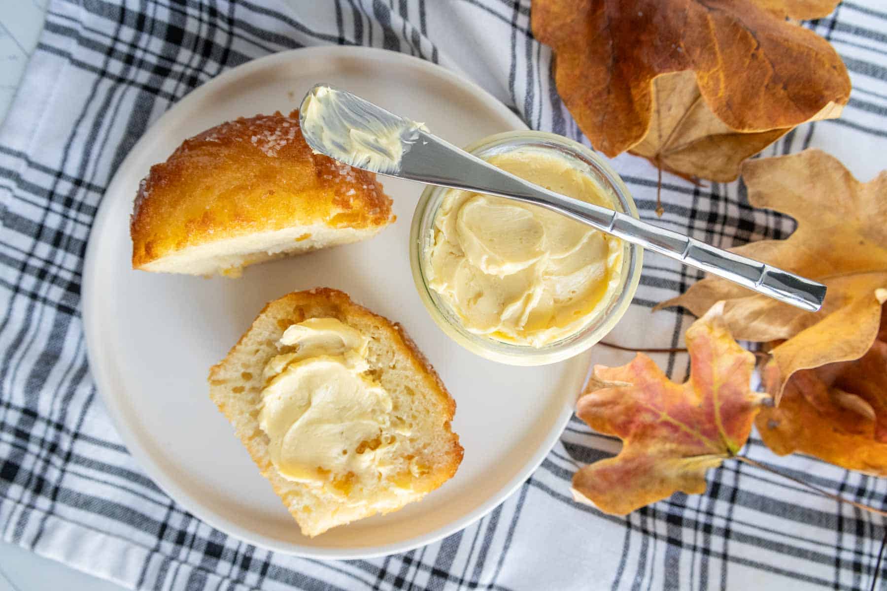 A plate with a sliced piece of bread spread with butter, a small jar of butter with a knife on top, and autumn leaves on a plaid cloth.