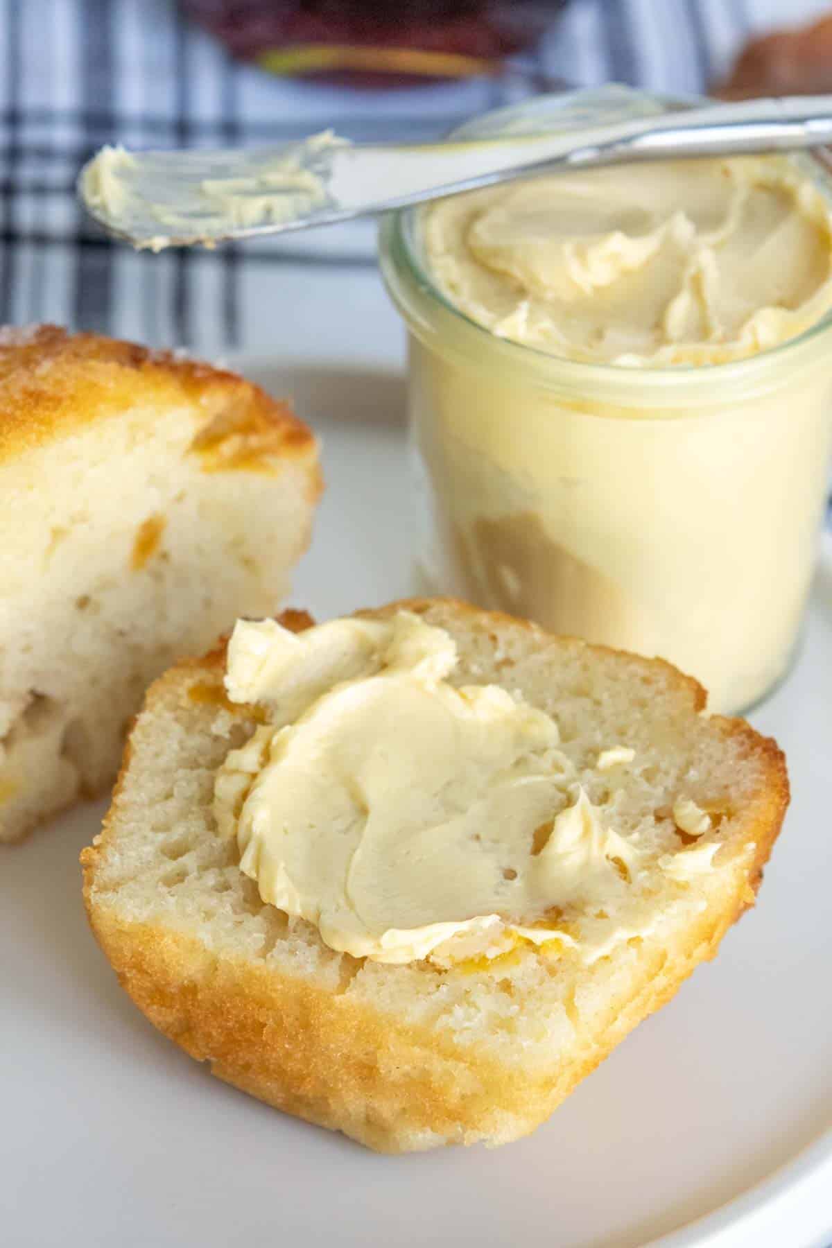 A muffin cut in half, spread with butter, next to a jar of butter with a buttered knife on top, on a white plate.