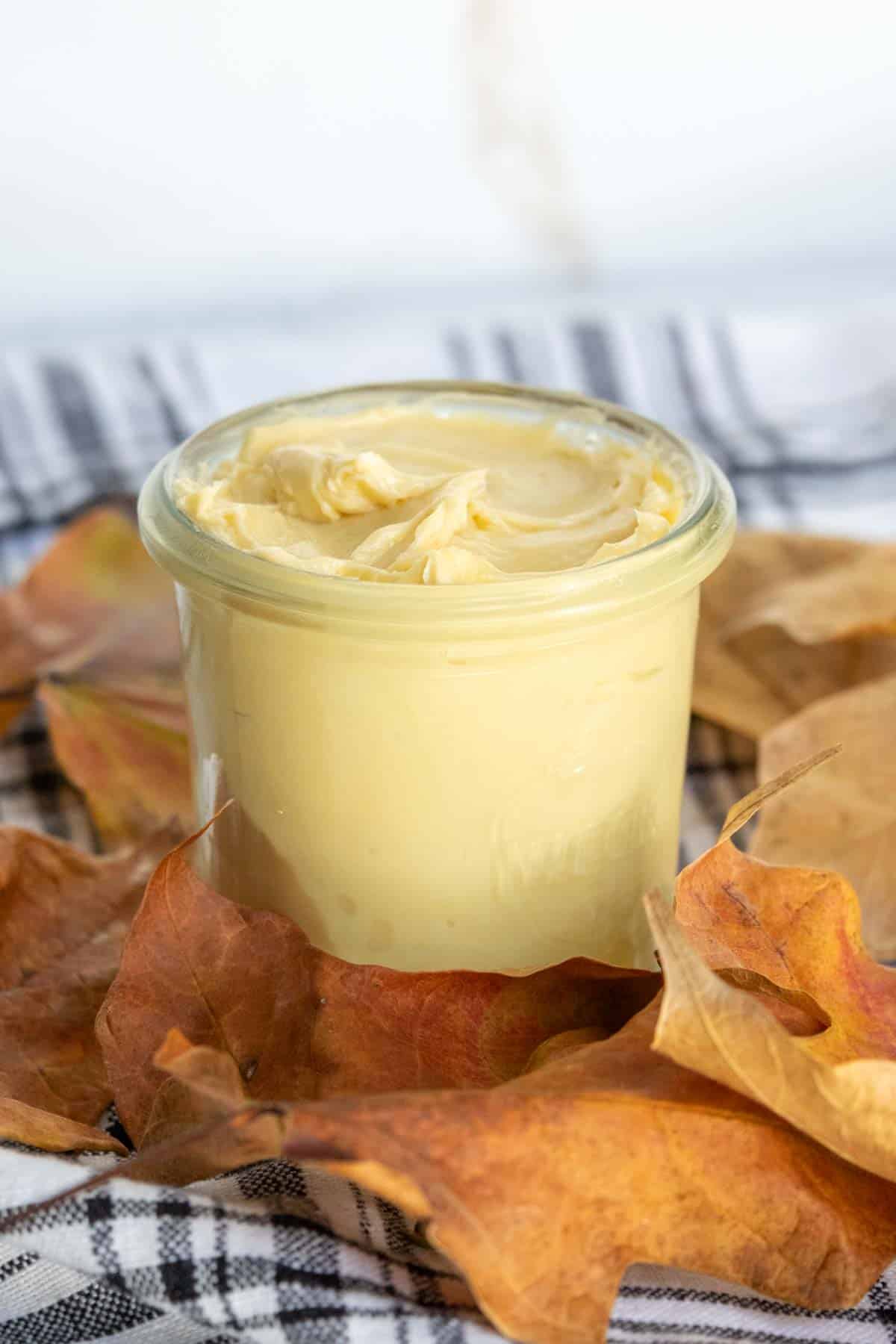 Glass jar filled with creamy yellow butter, surrounded by autumn leaves on a plaid fabric.