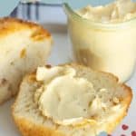 A halved biscuit spread with maple butter on a white plate, accompanied by a jar of the butter. Background features light blue text and red berry graphics.