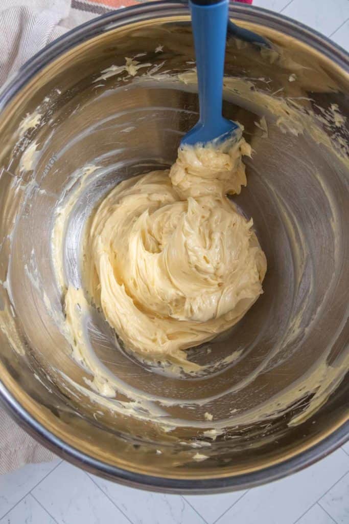 Metal mixing bowl with creamy yellow butter and a blue spatula resting inside.