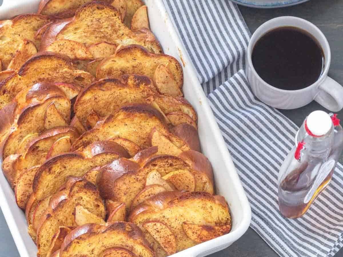 casserole dish of overnight apple cinnamon french toast with syrup and plates beside