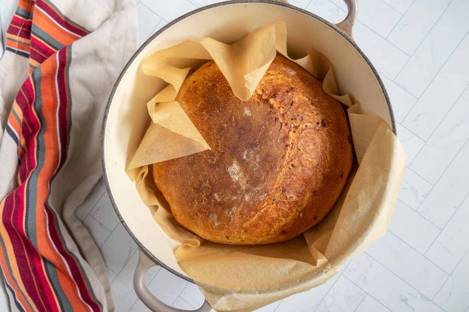 Round loaf of bread in a parchment-lined pot. A striped towel is on the side.