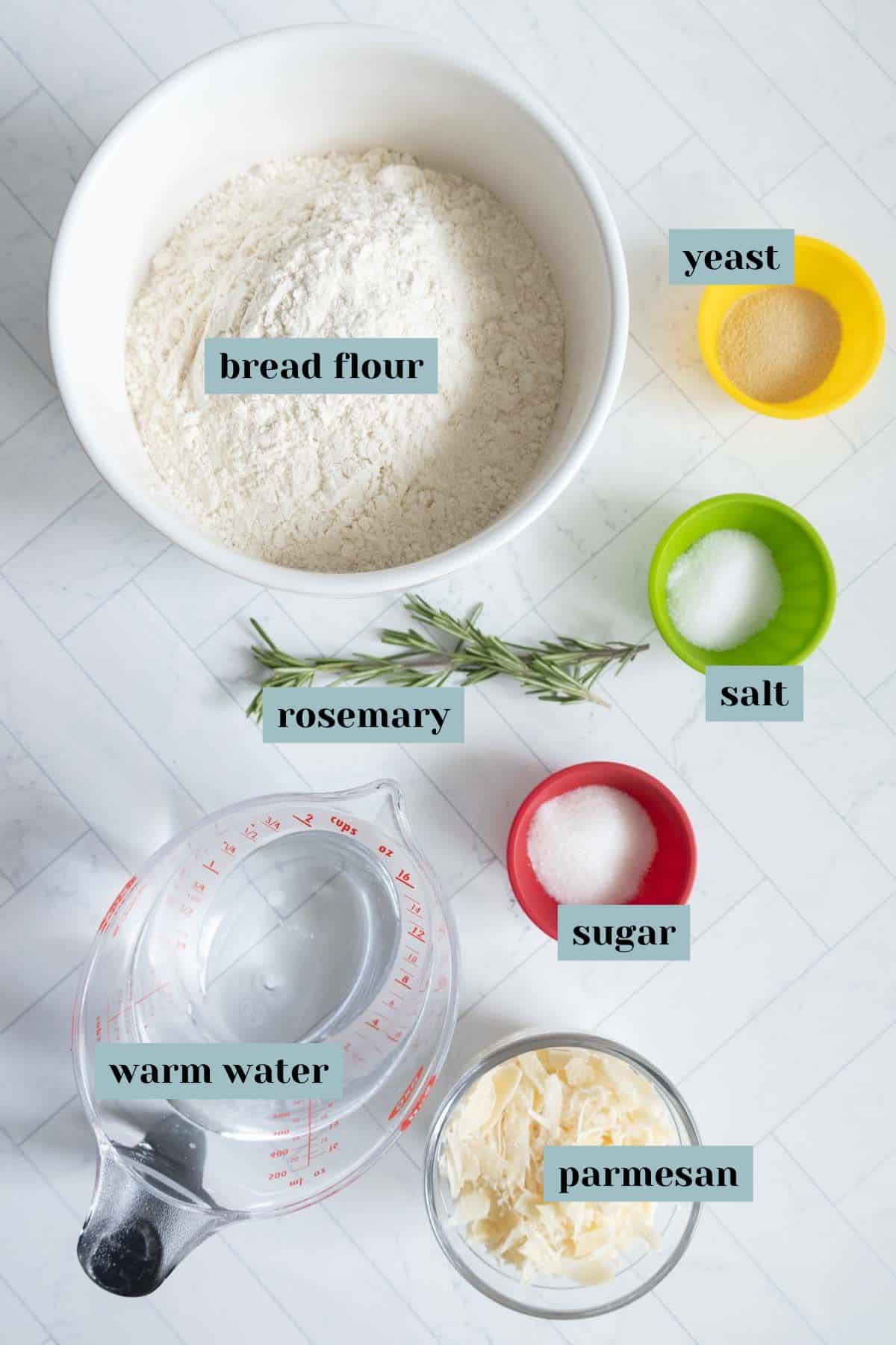 Ingredients for bread: bowl of bread flour, yeast in a yellow bowl, salt in a green bowl, sugar in a red bowl, parmesan in a white dish, rosemary sprigs, and a measuring cup with warm water.