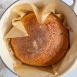 A round loaf of crusty bread in a parchment-lined pot, viewed from above on a tiled surface.