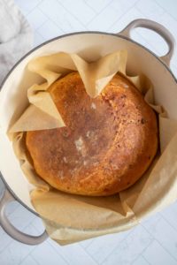 A round loaf of crusty bread in a parchment-lined pot, viewed from above on a tiled surface.