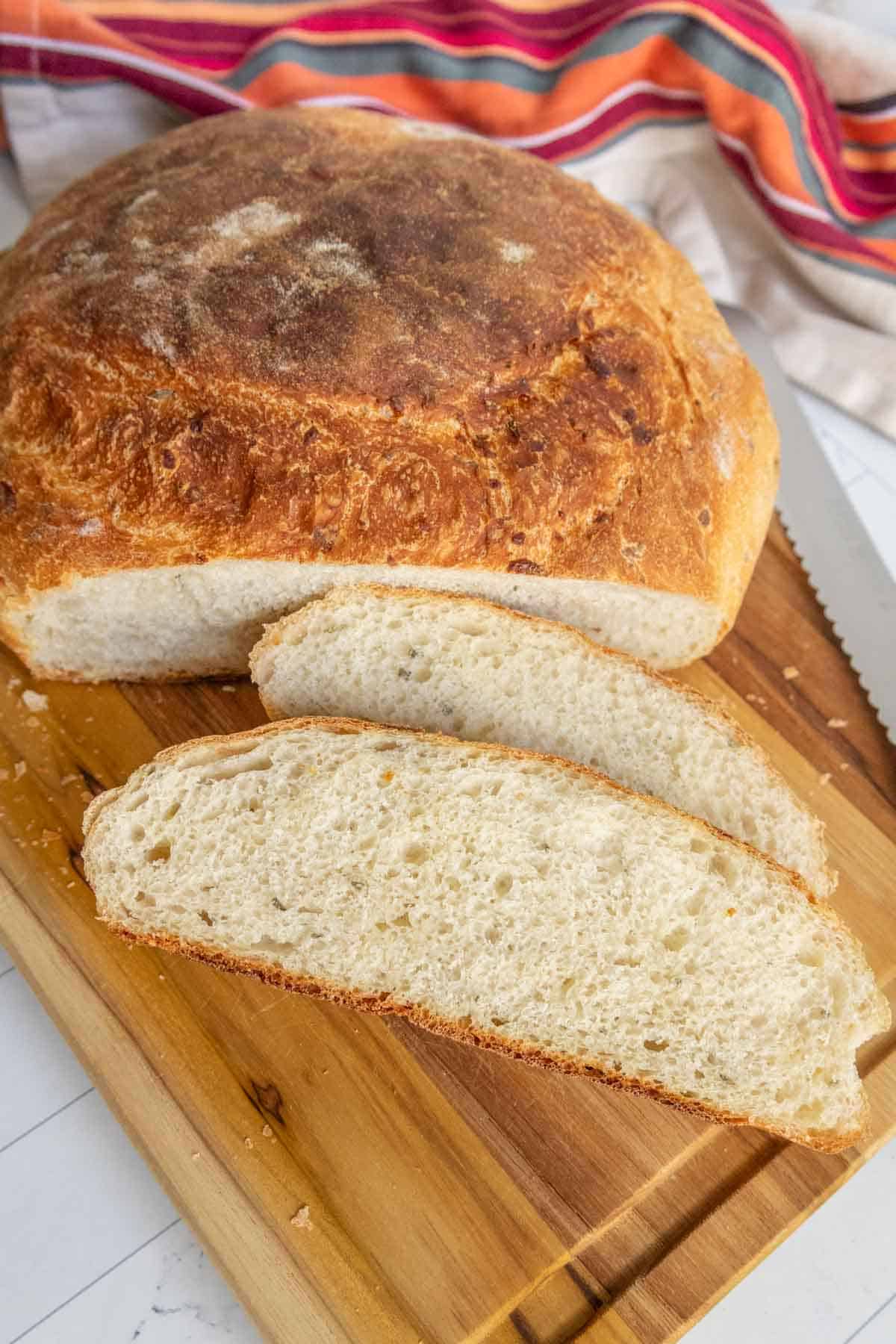 A loaf of crusty bread with two sliced pieces on a wooden cutting board. A serrated knife and striped cloth are nearby.