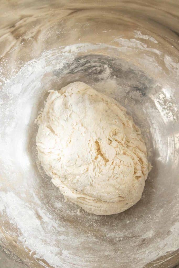 A ball of dough in a floured metal bowl.