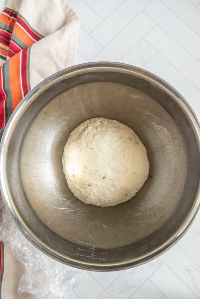 A ball of dough rests in a metal mixing bowl on a tiled surface, next to a striped cloth.