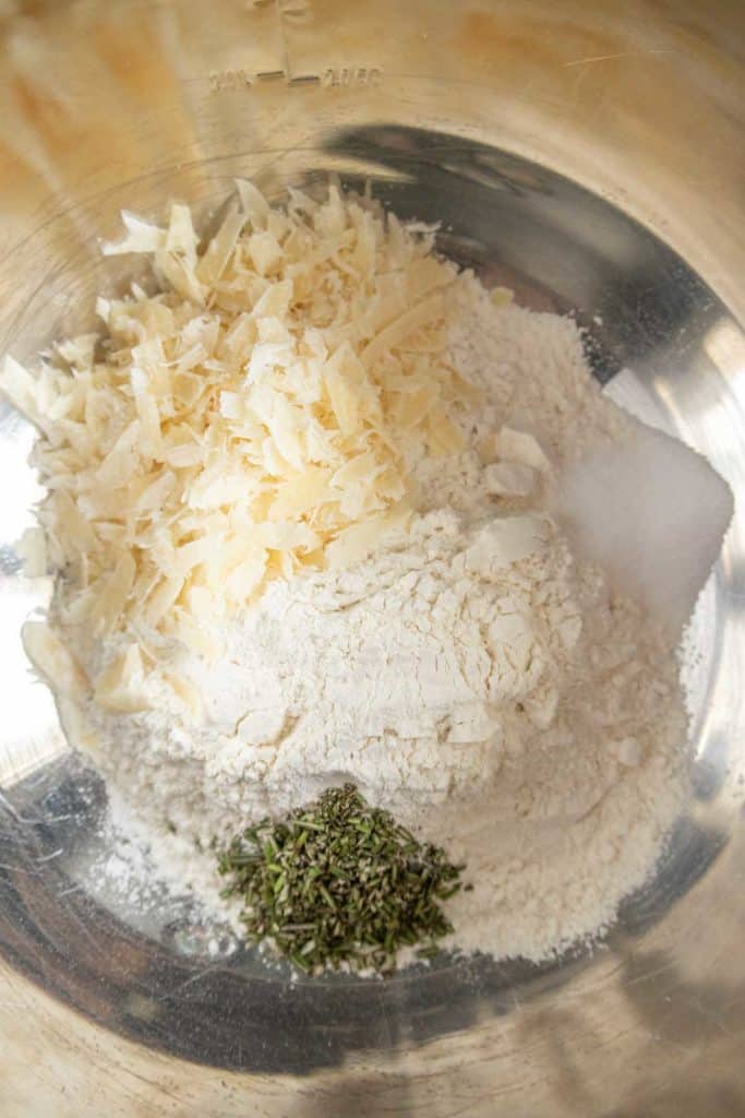 A bowl contains shredded cheese, flour, salt, and fresh herbs ready for mixing.