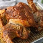 Roasted chicken on a cooling rack with crispy, golden skin, garnished with fresh herbs, placed on a light-colored surface.