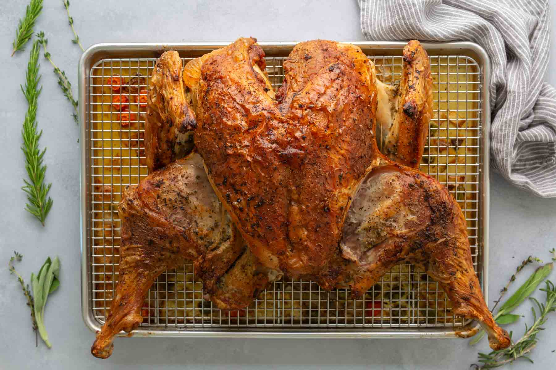 Roasted spatchcock turkey on a wire rack over a baking sheet, surrounded by herbs and a striped cloth.