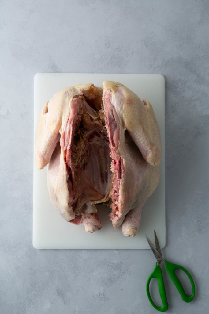 A raw, spatchcocked chicken on a cutting board next to green-handled kitchen scissors.